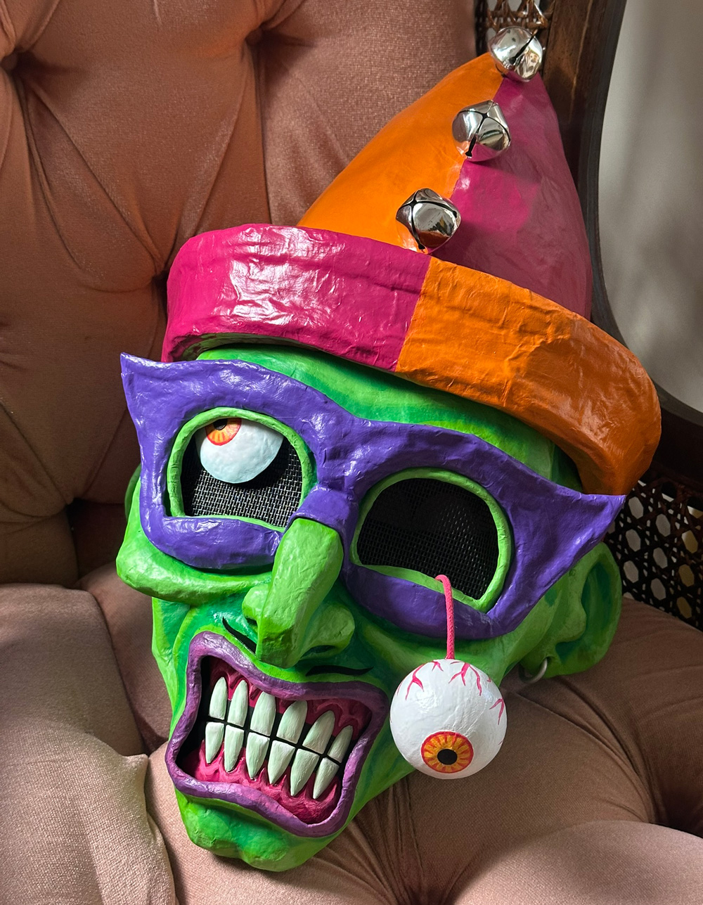 The completed Fortunato mask with a green face, dangling eyeball, and orange and magenta jester's cap