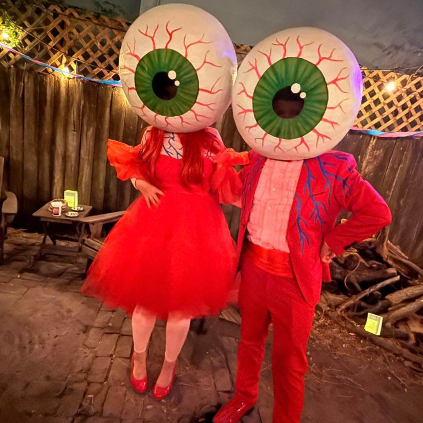 Two people wearing giant eyeball masks over their heads. The lady is wearing a red dress and the guy is wearing a red suit.