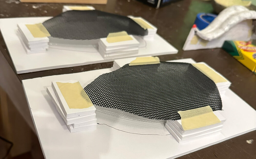 Two irregular-shaped pieces of black screen material taped onto foam supports for painting.