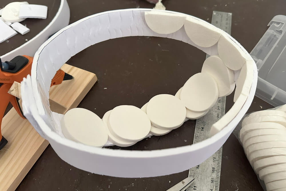Upside-down headpiece structure; a headband-shaped strip of foam board with several white makeup pads glued inside, with another strip as the crest, also with makeup pads glued into it.