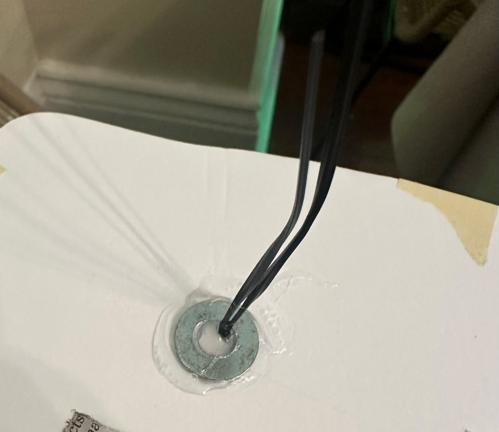 A metal washer with a twist tie sticking out of it, hot-glued to a panel of foam board.