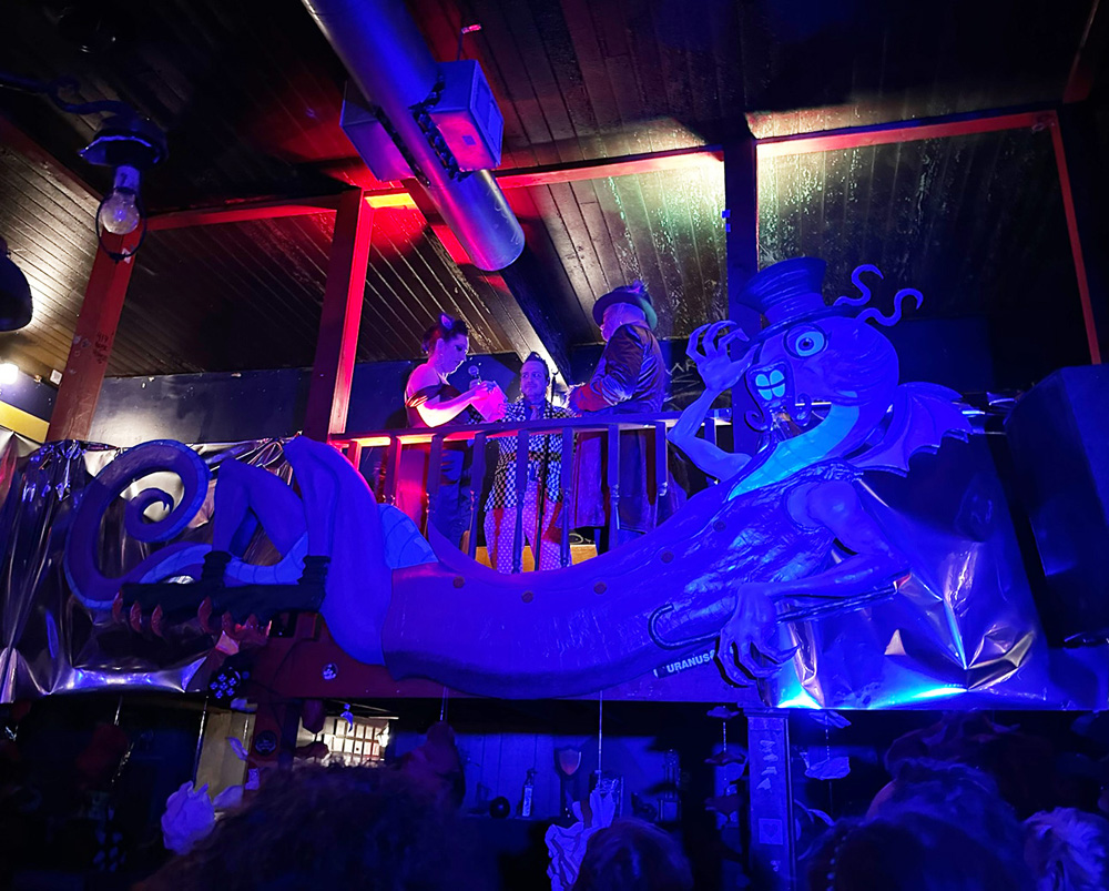 The Jabberwocky sculpture hanging along the upstairs railing of a bar in bright blue/purple light during the wedding. The lovely couple are visible above it.