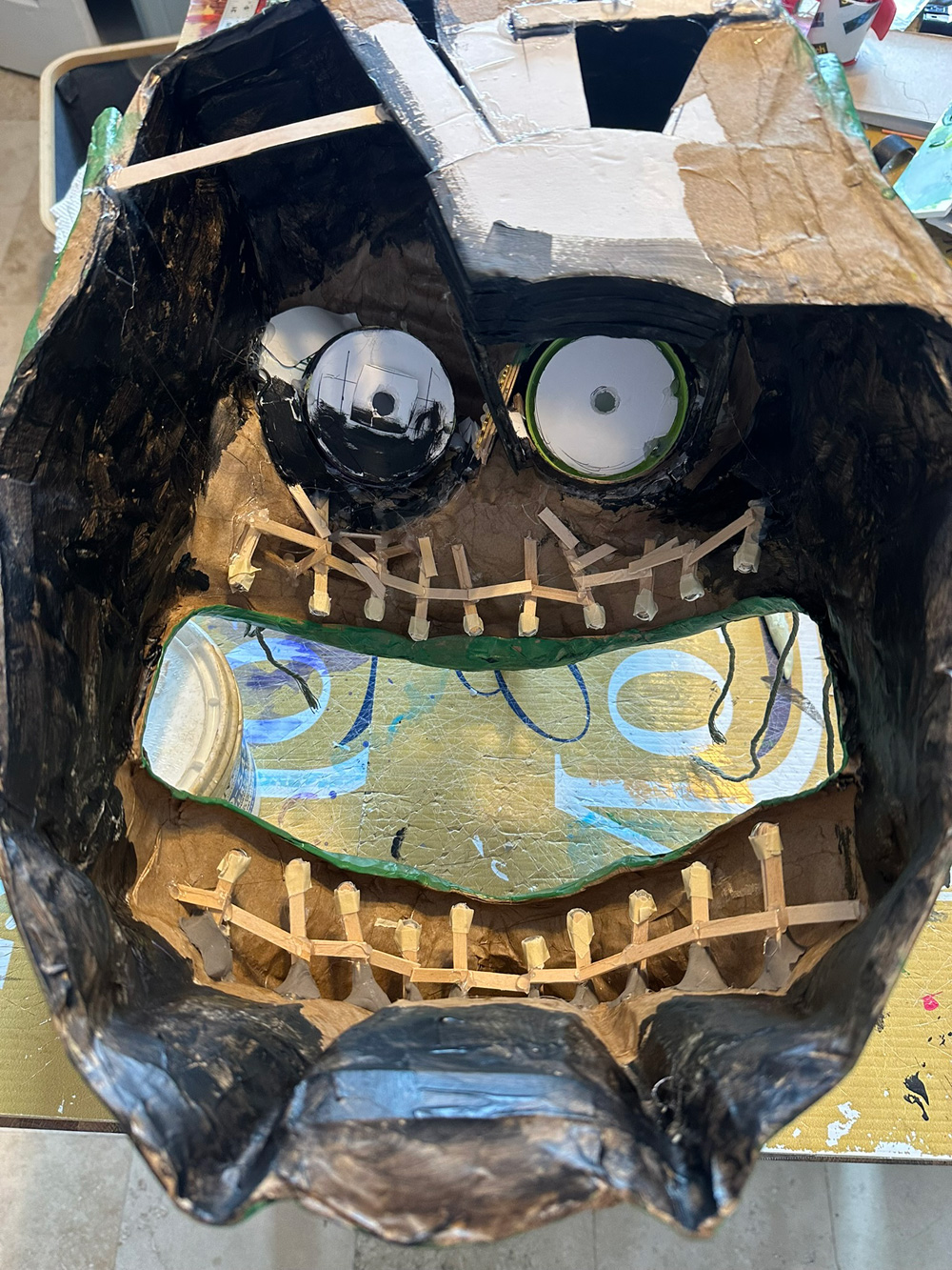 Interior view of the witch head from the back; it looks like a mess. Where the eyes are, there are white foam board disks with holes in them; this is where the lights will be inserted. In the mouth, there are twenty little structures made from wooden coffee stirrers hot-glued together; these are the areas where the teeth will be attached with magnets. Most of the interior of the head is painted black.